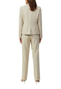 Costume beige avec veste ajustée et pantalon à jambe droite. Tissu lisse, design minimal, et détail de pli au dos de la veste. Sandales à talons présentées.