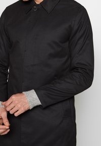 Muž oblečený v černém zapínacím kabátu s šedým pleteným svetrem pod ním, upravující rukáv, přičemž jsou vidět obě ruce.