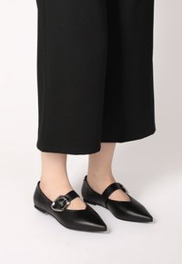 Černé kožené baleríny s špičatou špičkou, páskem přes nárt a dekorativní metalickou přezkou. Matný povrch a nízký podpatek.