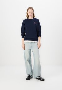 Sudadera navy con cuello redondo y puños de punto, combinada con jeans de pierna ancha en azul claro. Modelo usando zapatos negros y una camiseta interior blanca.