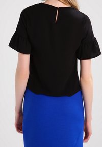 Femme portant un haut noir à manches courtes avec des manches volantées et un dos à encolure clé, assorti à une jupe bleu vif.