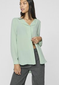 Blusa verde chiaro in un tessuto morbido e fluente. Presenta un colletto a punta, chiusura con bottoni e maniche lunghe con polsini bottonati. Vestibilità ampia.