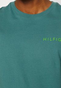 Nærbillede af en person iført en turkist crew-neck t-shirt med grøn "HILFIGER" tekst på brystet.