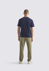 Man som står med ryggen mot kameran, iklädd en marinblå t-shirt, khakibyxor och vita sneakers mot en ljus, enfärgad bakgrund.