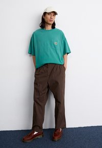 T-shirt turcoaz cu buzunar, pantaloni maro lejeri și mocasini maro cu ciucuri. Detalii de cusătură îngrijite și culori contrastante. Include o șapcă neutră.