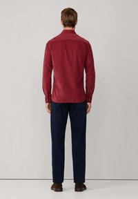 Rode corduroy overhemd met een button-down kraag, zichtbare naden en knoopmanchetten, gecombineerd met marineblauwe broek en bruine schoenen, gezien van achteren.