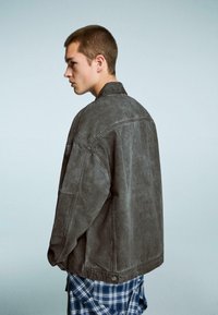 Siva prevelika denim jakna z teksturiranim finišem, vidnim šivanjem in ovratnikom. Nosi se preko modre karirane srajce.