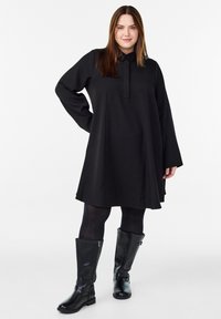 Žena stojící sebevědomě v volných černých šatech, černých punčochách a vysokých černých kožených botách na pozadí bílé barvy.