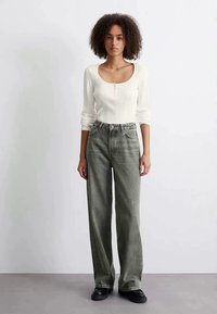 Mulher com cabelo encaracolado, vestindo uma blusa branca de manga longa em renda e jeans cinzentos folgados, de pé contra uma parede branca lisa.