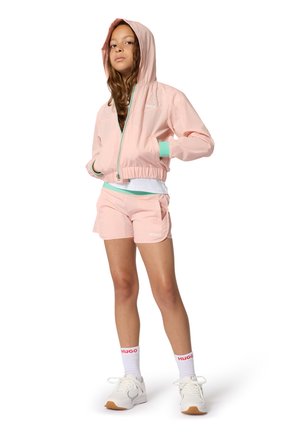 Mädchen in einer pinkfarbenen Kapuzenjacke und Shorts, dazu weiße HUGO-Socken und Sneakers, steht mit den Händen in den Jackentaschen.