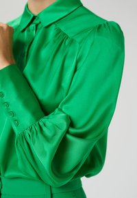 Blusa in satin verde con colletto, maniche a sbuffo e dettagli con bottoni sui polsini. Texture liscia con una finitura brillante.