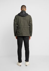 Olivgrön jacka med en svart, vadderad huva, med en ficka på ryggen och åtsittande ärmslut. Bärs över svarta byxor och skor.