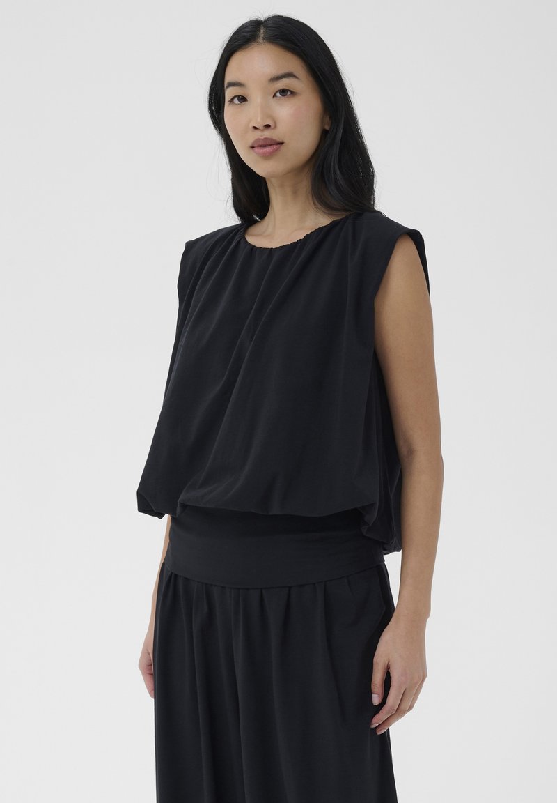 Femme aux longs cheveux noirs portant un haut noir sans manches et ample ainsi qu'une jupe noire assortie, debout devant un fond clair uni.
