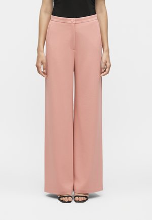 Femme portant un pantalon rose taille haute à jambes larges avec bouton à l’avant et des talons ouverts noirs, debout devant un fond uni.