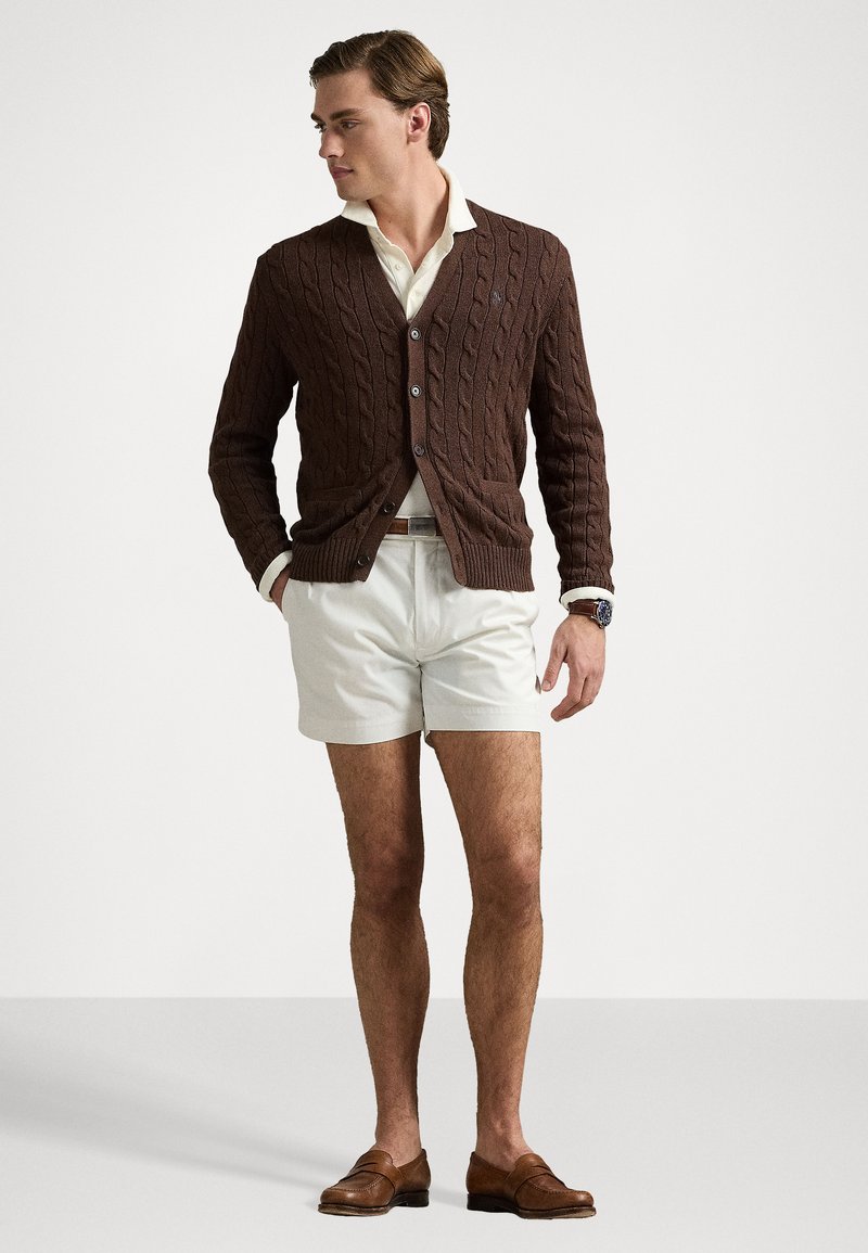 Cardigan marrone a trecce con bottoni, indossato sopra una camicia bianca con colletto e pantaloni corti chiari. Mocassini marroni completano l'outfit.