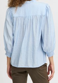Blouse bleu clair avec des manches bouffantes et un dos froncé. Présente un tissu texturé et des poignets smockés. Design décontracté adapté à diverses occasions.