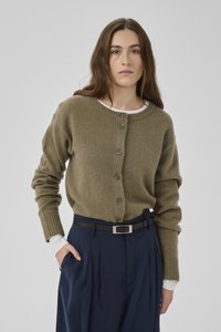 Junge Frau mit langen braunen Haaren, die eine olivgrüne, geknöpfte Strickjacke über einem weißen Oberteil und marineblaue, mit Gürtel versehene Hose trägt, vor grauem Hintergrund.