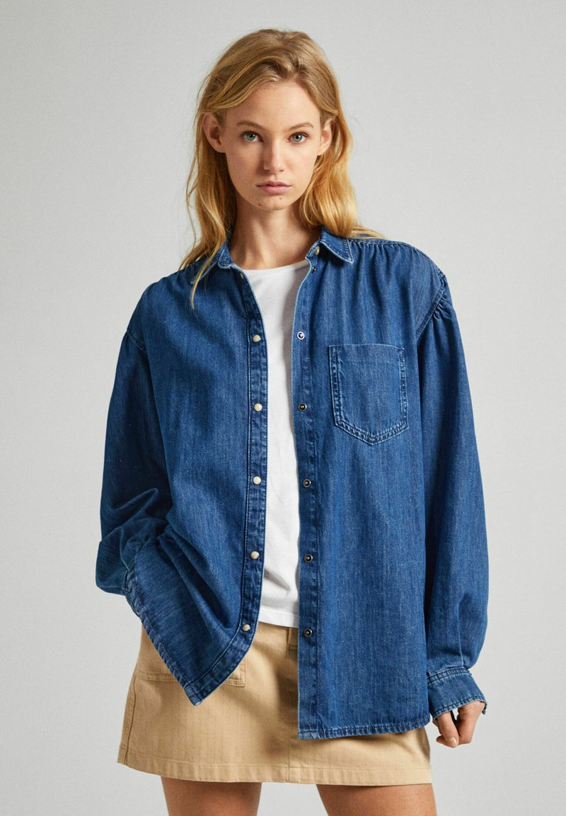 Camicia di denim con una chiusura a bottoni frontale, maniche lunghe, taschino sul petto e una vestibilità comoda. Indossata sopra una maglietta bianca e abbinata a una gonna beige.