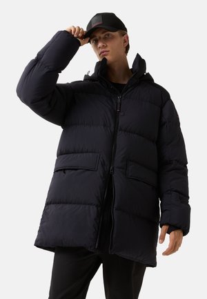 Sort pufferjakke med hætte, der har lynlås foran, sidelommer og quiltet tekstur. Bæres med en sort kasket, mens modellen poserer.