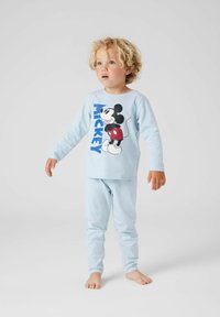 Malé dieťa s kučeravými blond vlasmi, oblečené v svetlomodrých pyžamkách s Mickey Mouse, stojí naboso na jednolatom bielom pozadí.
