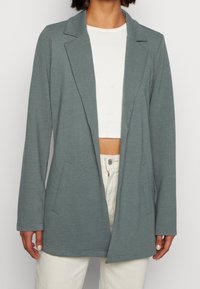 Blazer grigio a coste con rever a lancia, apertura frontale e maniche lunghe. Presenta due tasche frontali e una forma leggermente oversize.