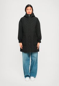 Vrouw in een zwarte, doorgestikte, capuchonjas met vóórzakken, blauwe wijde jeans en witte sneakers, staande tegen een effen lichte achtergrond.
