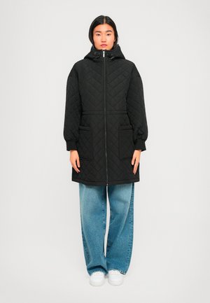 Woman wearing a black quilted hooded coat with front pockets, blue wide-leg jeans, and white sneakers, standing against a plain light background.