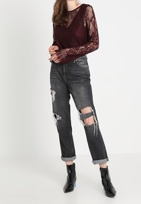 Bordeaux blonde langærmet top med dyb V-hals og gennemsigtige ærmer, parret med slidte sorte jeans med revner ved knæene og opsmøgede cuffs.