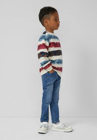 Malý chlapec stojaci bokom, oblečený v pruhovanom, viacfarebnom svetri, modrých džínsoch a bielych teniskách, s rukami v vreckách na pozadí bez vzoru.