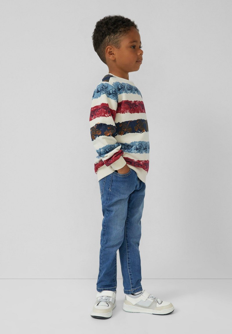 Malý chlapec stojaci bokom, oblečený v pruhovanom, viacfarebnom svetri, modrých džínsoch a bielych teniskách, s rukami v vreckách na pozadí bez vzoru.