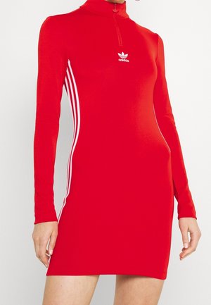Rode Adidas jurk met lange mouwen, witte drie strepen aan de zijkanten, ritskraag en logo op de borst, gedragen door een persoon die met ontspannen armen staat.