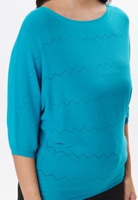 Maglione turchese lavorato a maglia con scollatura larga, maniche corte e motivo a zigzag nella lavorazione aperta sulla parte anteriore. Texture morbida e vestibilità rilassata.