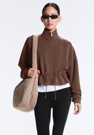 Frau trägt braune Jacke mit Reißverschluss und schwarze Sonnenbrille, trägt eine große beige Umhängetasche, steht vor einfarbigem Hintergrund.