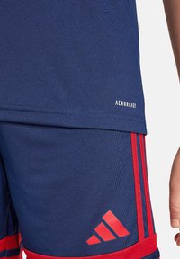 Marineblauw sportshirt met rode strepen en "AEROREADY" tekst. Glad oppervlak met vochtafvoerende stof; logo op de shorts in een rode accent.