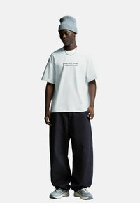 Jongen in een grijze beanie, witte T-shirt met tekst, zwarte wijde broek, zilveren ketting, en grijze sneakers, staande en naar beneden kijkend.