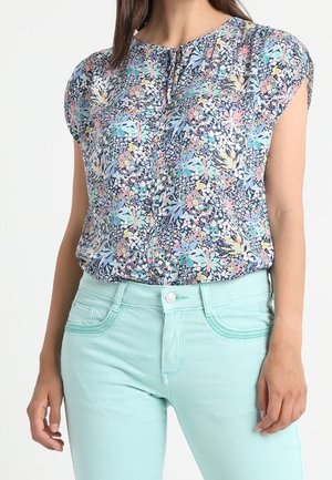 Kvinde iført en kortærmet blomstret bluse med pastelfarvede blå-grønne tætsiddende bukser, stående foran en ensfarvet hvid baggrund.