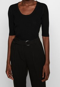 Musta ribbed paita, jossa on avara kaula-aukko ja lyhyet hihat, yhdistettynä korkeavyötäröisiin mustiin housuihin, joissa on vyö ja metallinen solkiyksityiskohta.