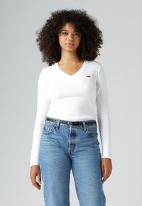 Bijela košulja dugih rukava s V-izrezom i malim crvenim Levi's logotipom, u kombinaciji s plavim trapericama visokog struka i crnim pojasom.