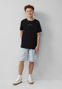 Zwarte katoenen T-shirt met "Sophie Club" in het wit gedrukt, gecombineerd met lichtblauwe denim shorts en witte sneakers met rubberen zolen.