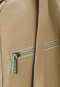 Sac en cuir beige texturé avec une sangle verticale visible et une fermeture éclair horizontale munie d'un curseur argenté gravé de la marque.