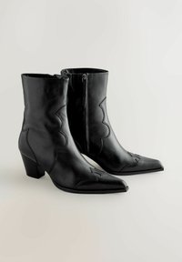 Par de botas de cuero negro hasta la mitad de la pantorrilla con tacones cuadrados, puntas afiladas, costuras decorativas y cremalleras laterales sobre un fondo claro.