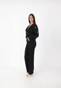 Svart cardigan med vita hjärtaccent, ihopparad med högmidjade svarta byxor. Modellen bär strappy vita klackar. Minimalistisk bakgrund.