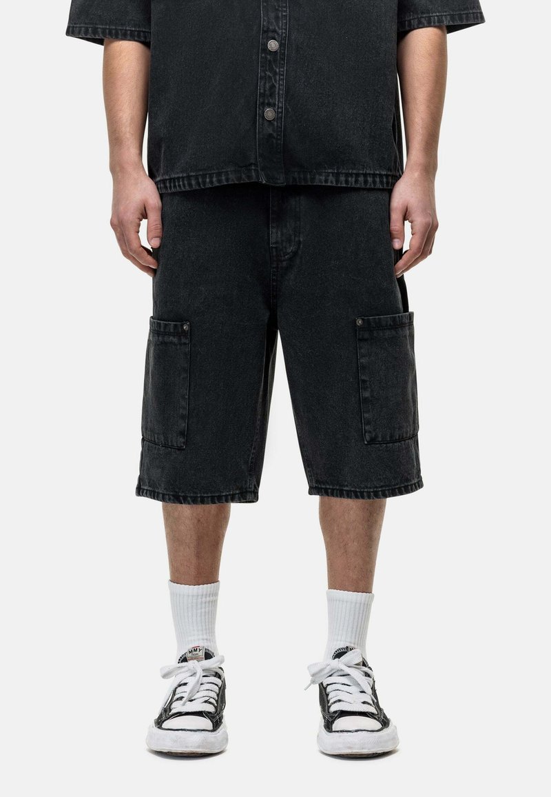 Shorts cargo en denim noir avec deux poches avant, présentant un ourlet brut et une coupe relâchée. Associés à des baskets noires et blanches et des chaussettes blanches.