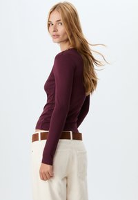 Langærmet maroon top i en tætsiddende design, lavet af blødt stof, parret med højhalset cremede bukser og et brunt bælte.