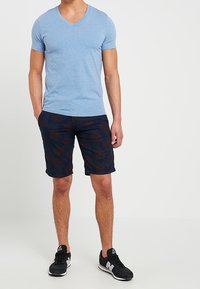 Maglietta blu chiaro a scollo a v abbinata a pantaloni corti scuri con fantasia e scarpe da ginnastica nere, che mette in evidenza un design aderente e uno stile casual.