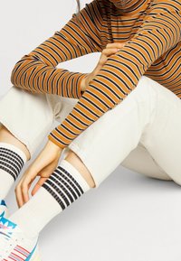 Camiseta de manga larga a rayas en naranja y negro, pantalones blancos y calcetas hasta la rodilla con un patrón de rayas negras. Las zapatillas tienen detalles en azul y rojo.