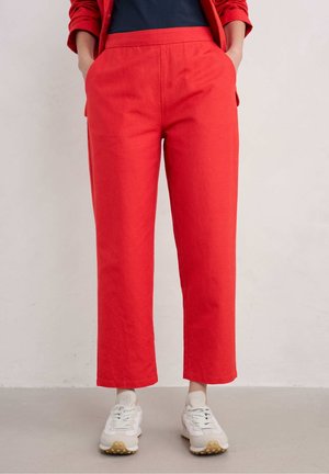 Henkilö, jolla on kirkkaanpunaiset suorat housut, musta paita ja valkoiset lenkkarit, seisoo käsivarsien taskuissa yksiväristä taustaa vasten.