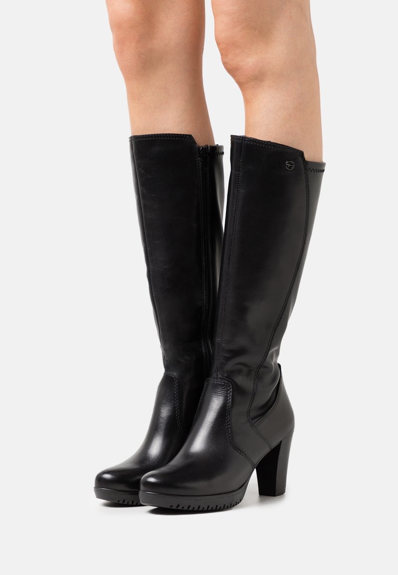 Bottes en cuir noires montantes jusqu'aux genoux avec fermetures éclair latérales et talons carrés portées sur jambes nues sur un fond clair uni.