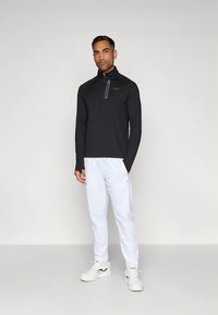 Pulover negru cu mâneci lungi și fermoar până la piept, combinat cu pantaloni sport albi. Pantofii sunt albi cu accente negre.