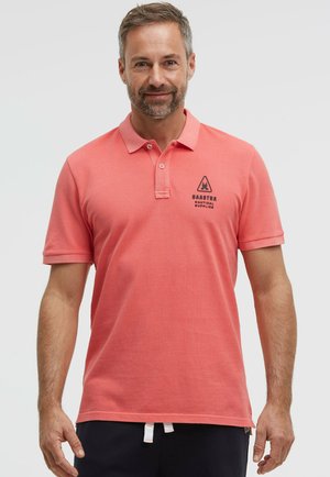 Mann trägt ein korallenrosafarbenes Polohemd mit kurzen Ärmeln und einem "Gaastra Nautical Supplies"-Logo auf der linken Brust vor einem einfarbigen Hintergrund.